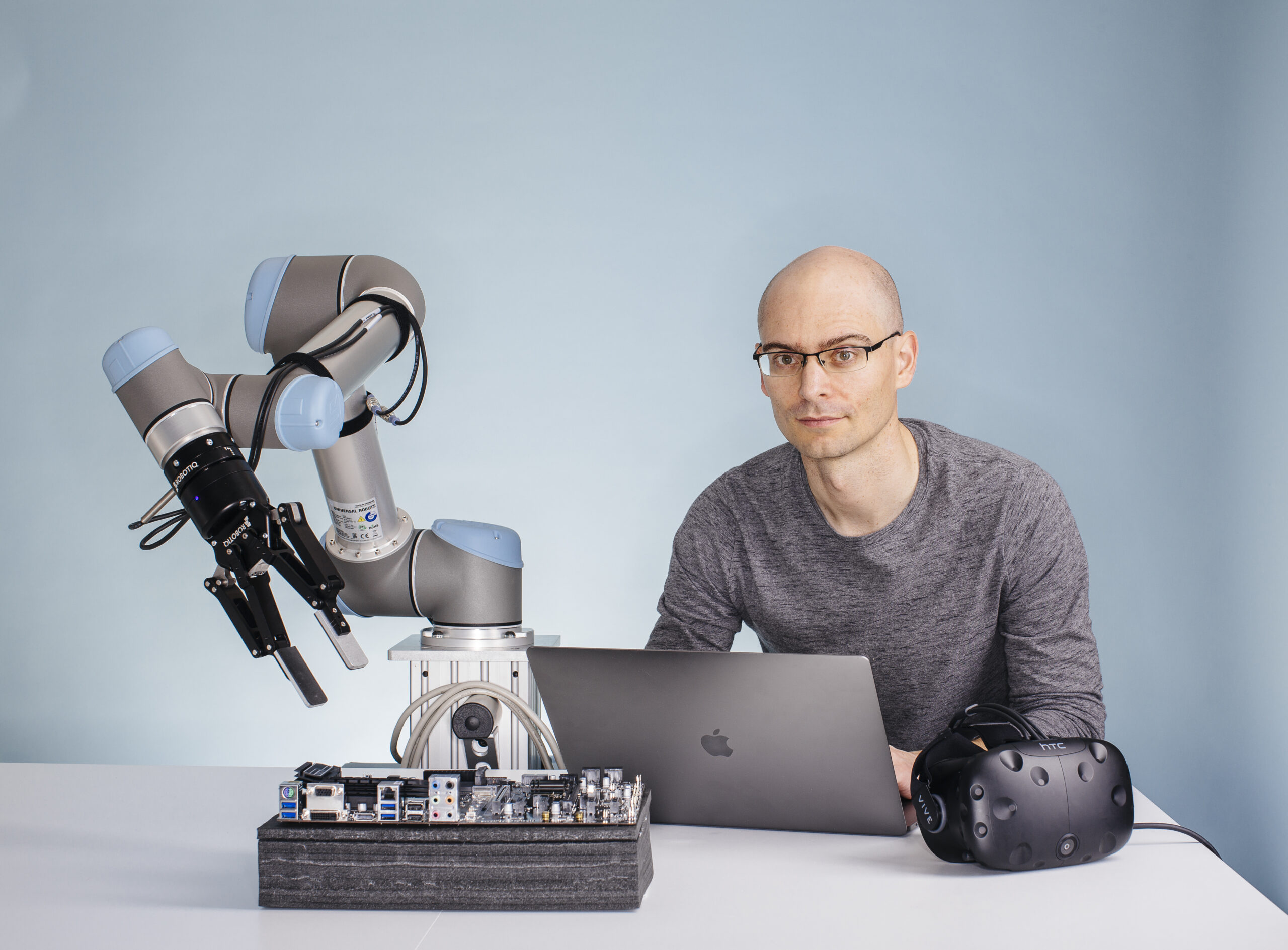 Pieter Abbeel posing in front of a blue background with a robot on desk